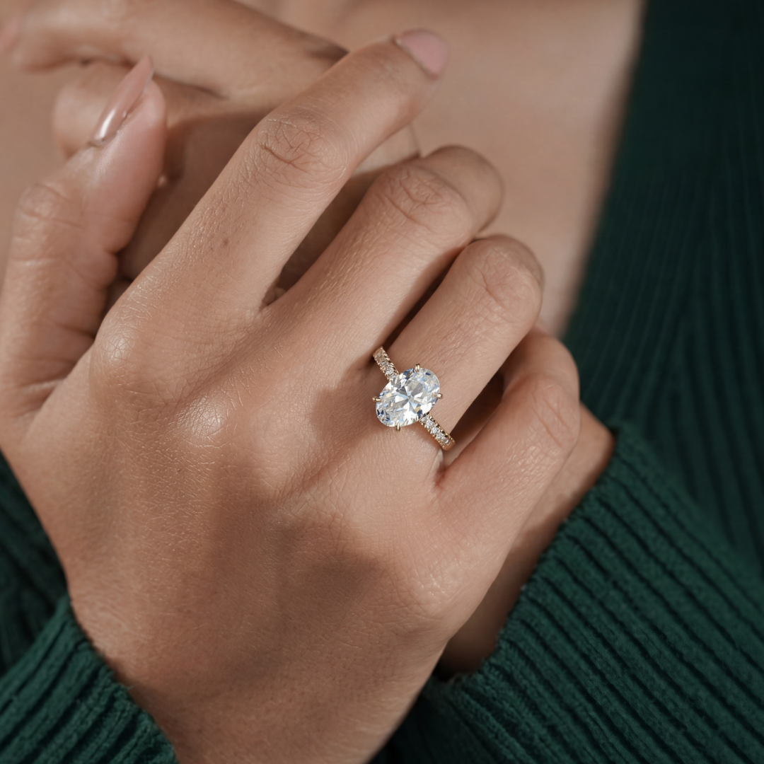 A close-up of a person's hand wearing a yellow-gold engagement ring with a large oval-cut clear center stone and small side stones, set against a dark green ribbed sweater.