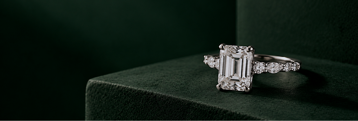 An emerald-cut diamond engagement ring with a white-gold band and side diamonds, displayed on a dark green velvet surface.